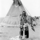 Historische Schwarz-Weiß-Fotografie: Ein Mann in traditioneller indigener Kleidung mit Perlenstickerei steht vor einem Tipi und hält einen Federfächer in der Hand