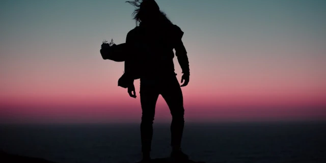 Silhouette eines Menschen mit wehenden Haaren auf einem Felsvorsprung über dem Meer bei Sonnenuntergang – Sinnbild für das Erreichen der Spitze