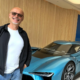 Andreas Schönemann steht lächelnd vor einem futuristischen blauen Elektro-Concept-Car in einem Showroom mit Holzverkleidung.
