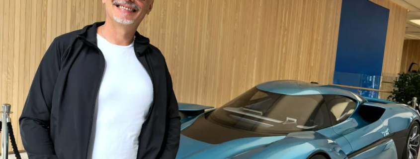 Andreas Schönemann steht lächelnd vor einem futuristischen blauen Elektro-Concept-Car in einem Showroom mit Holzverkleidung.